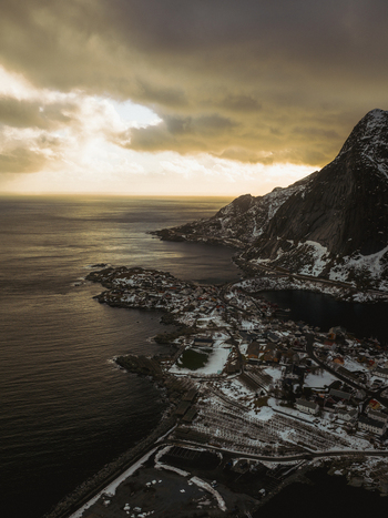Aerial view of Reinebringen s snow dusted slopes meet the moody Atlantic under a sky brushed with gold and grey Reinebringen Nordland Norway.
