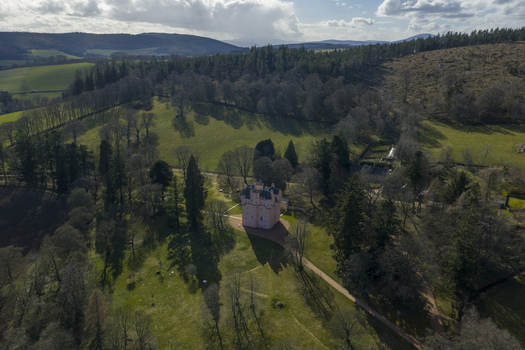 Aerial view of the iconic pink Craigievar Castle nestled among lush green fields and dense woodland under a dramatic sky in Craigievar Scotland United Kingdom.