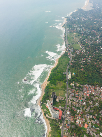Aerial view of the rugged coastline with waves crashing against the shore a coastal road and urban buildings surrounded by lush tropical greenery in Galle Southern Province Sri Lanka. Aerial view of the rugged coastline with waves crashing against the shore a coastal road and urban buildings surrounded by lush tropical greenery in Galle Southern Province Sri Lanka.