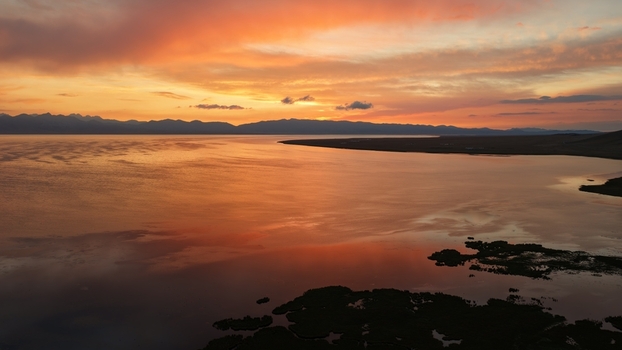 Aerial view of Song Kul lake at sunset with vibrant orange sky reflecting on the calm water and distant mountains in Naryn Region Kyrgyzstan. Aerial view of Song Kul lake at sunset with vibrant orange sky reflecting on the calm water and distant mountains in Naryn Region Kyrgyzstan.