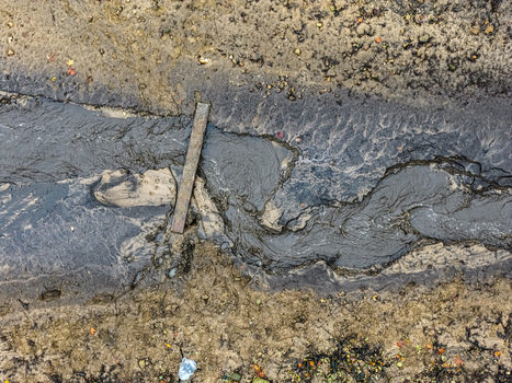 Aerial view of a muddy stream flowing through textured earth with a wooden plank crossing the water in Latvia.