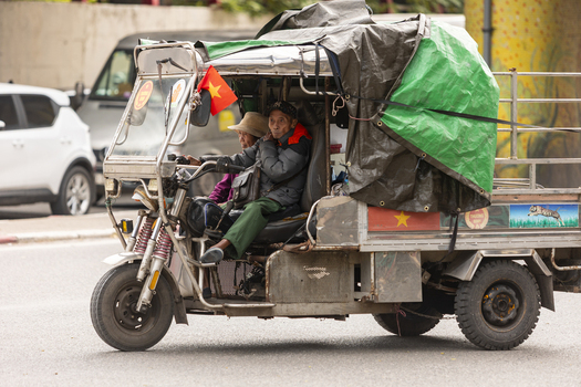 Hanoi Vietnam 11 November 2025 View of an elderly man driving a motorized three wheeled vehicle with a green tarp cover and a small Vietnamese flag on a city street.