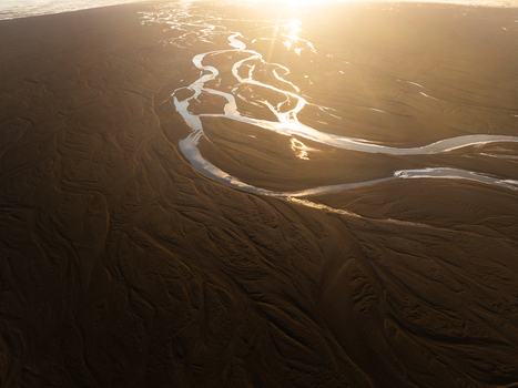 Aerial view of the braided river system near Lomagnupur with golden hour sunlight reflecting on the water and dark volcanic sand patterns across the vast landscape in Skaftarhreppur Iceland.
