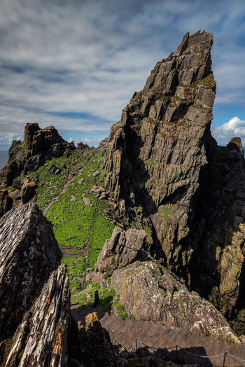 View of the jagged rock peaks and green slopes of Great Skellig with stone steps under a cloudy sky in County Kerry Ireland. View of the jagged rock peaks and green slopes of Great Skellig with stone steps under a cloudy sky in County Kerry Ireland.