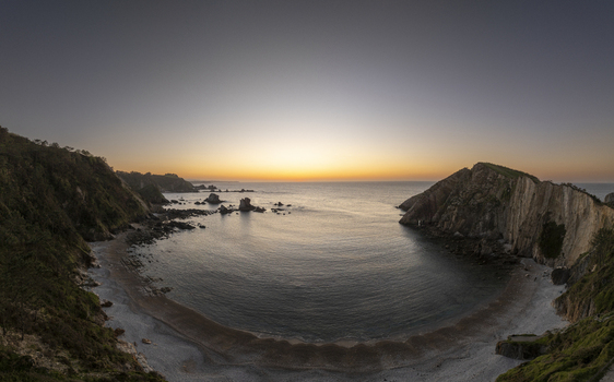 Aerial view of a serene coastal bay at sunset with rocky cliffs framing the sandy beach and calm waters in Nueva Asturias Spain.