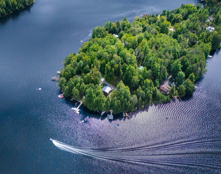 Aerial view of Baptiste Lake featuring a forested island with cottages docks and a motorboat creating a wake on the water in Hastings Highlands Ontario Canada.