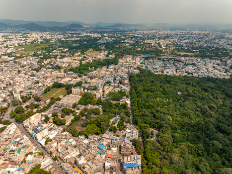 Aerial view of the urban landscape of Udaipur featuring dense residential buildings narrow streets and a large expanse of lush green forest in Udaipur Rajasthan India. Aerial view of the urban landscape of Udaipur featuring dense residential buildings narrow streets and a large expanse of lush green forest in Udaipur Rajasthan India.