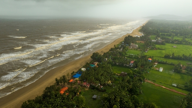 Aerial view of Gokarna beach with waves crashing on the sandy shore lush green palm trees and agricultural fields under a cloudy sky in Gokarna Karnataka India.