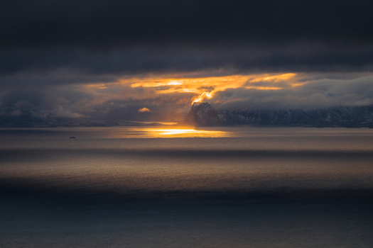 Aerial view of golden sunlight piercing through heavy clouds reflecting on the calm dark waters surrounding the distant snow capped mountains Henningsv&aelig;r Nordland Norway.