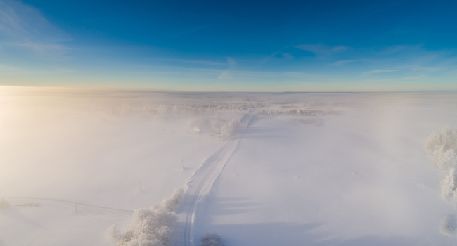 Aerial view of a snow covered road winding through a dense fog covered landscape with frost covered trees under a clear blue sky in Latvia. Aerial view of a snow covered road winding through a dense fog covered landscape with frost covered trees under a clear blue sky in Latvia.