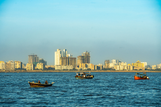 View of the Karachi skyline across the sea with traditional fishing boats carrying people in the foreground under a clear blue sky in Karachi Sindh Pakistan.