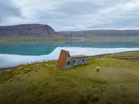 Aerial view of a lone figure in yellow standing near an abandoned house with a red roof reflected in the still fjord waters Westfjords Iceland. Aerial view of a lone figure in yellow standing near an abandoned house with a red roof reflected in the still fjord waters Westfjords Iceland.