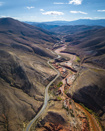 Aerial view of a winding road through a vast arid mountain valley with rugged slopes colorful rock formations and a dry riverbed under a bright blue sky in Argentina. Aerial view of a winding road through a vast arid mountain valley with rugged slopes colorful rock formations and a dry riverbed under a bright blue sky in Argentina.