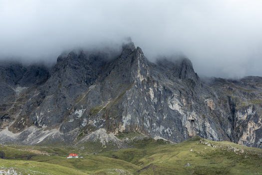 View of a solitary red roofed house nestled in the vibrant green foothills beneath towering jagged mountains shrouded in ethereal mist Picos de Europa Asturias Spain. View of a solitary red roofed house nestled in the vibrant green foothills beneath towering jagged mountains shrouded in ethereal mist Picos de Europa Asturias Spain.