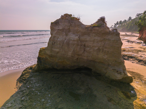 Aerial view of a large rock formation on a sandy beach with ocean waves and palm trees along the coastline in Bali Indonesia. Aerial view of a large rock formation on a sandy beach with ocean waves and palm trees along the coastline in Bali Indonesia.