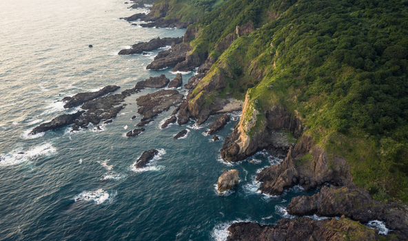 Aerial view of the rugged coastline featuring steep rocky cliffs lush green forests and the deep blue sea with crashing waves in Izu Shizuoka Japan. Aerial view of the rugged coastline featuring steep rocky cliffs lush green forests and the deep blue sea with crashing waves in Izu Shizuoka Japan.