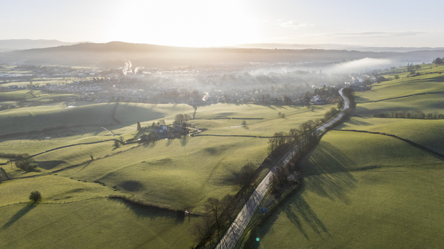 Aerial view of a winding road cutting through vibrant green fields with a distant town shrouded in soft morning mist England United Kingdom. Aerial view of a winding road cutting through vibrant green fields with a distant town shrouded in soft morning mist England United Kingdom.