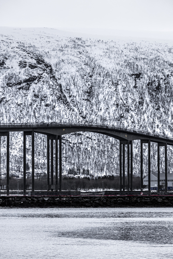 View of the Tromso Bridge with its concrete pillars and arched span against a backdrop of snow covered mountains Tromso Troms Norway. View of the Tromso Bridge with its concrete pillars and arched span against a backdrop of snow covered mountains Tromso Troms Norway.
