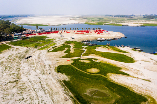 Bogura Bangladesh 06 March 2022 Aerial view of red fabrics drying on the sandy riverbank with boats moored in the water and green fields nearby.