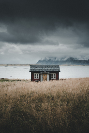 View of a traditional red wooden cabin situated in a field of dry grass overlooking a calm fjord with distant mountains under a dramatic overcast sky in Lofoten Nordland Norway. View of a traditional red wooden cabin situated in a field of dry grass overlooking a calm fjord with distant mountains under a dramatic overcast sky in Lofoten Nordland Norway.