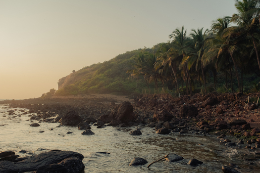 View of rocky shorelines meet lush green hills dotted with palm trees under a soft hazy sky bathed in the warm glow of the setting sun Chiplun Maharashtra India. View of rocky shorelines meet lush green hills dotted with palm trees under a soft hazy sky bathed in the warm glow of the setting sun Chiplun Maharashtra India.