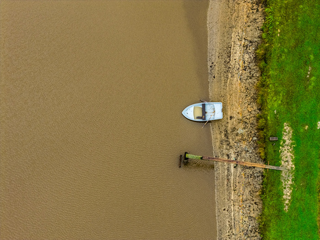 Aerial view of a small boat moored along a riverbank with a wooden pier and green grassy edge in Latvia.
