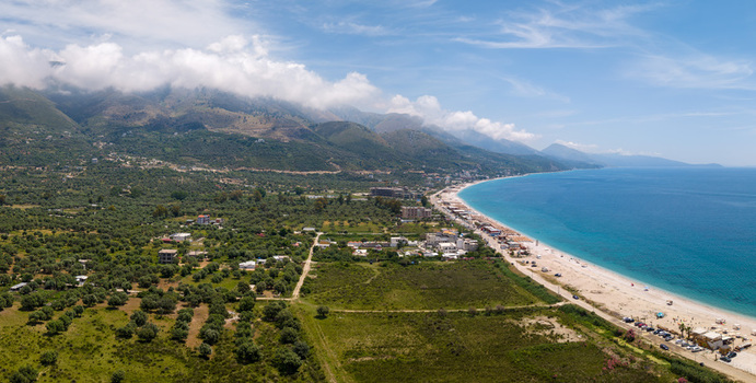 Aerial view of the turquoise coastline and sandy beach of Borsh surrounded by lush olive groves and majestic mountains under a blue sky in Borsh Vlore County Albania. Aerial view of the turquoise coastline and sandy beach of Borsh surrounded by lush olive groves and majestic mountains under a blue sky in Borsh Vlore County Albania.