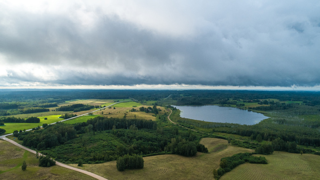 Aerial view of a serene rural landscape featuring a tranquil lake surrounded by lush green forests rolling fields and a winding road under a dramatic cloudy sky Latvia. Aerial view of a serene rural landscape featuring a tranquil lake surrounded by lush green forests rolling fields and a winding road under a dramatic cloudy sky Latvia.