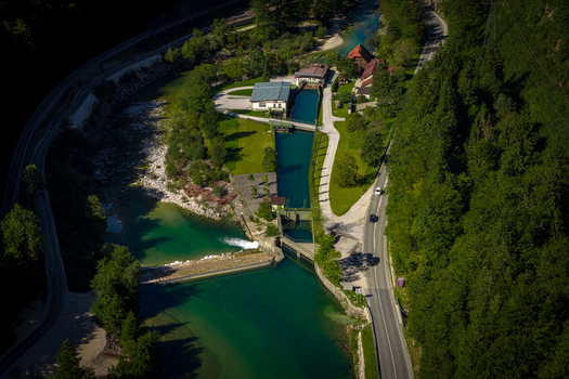 Aerial view of the Sava Bohinjka river featuring a hydroelectric power plant weir and surrounding dense forest with a road running alongside in Radovljica Slovenia.