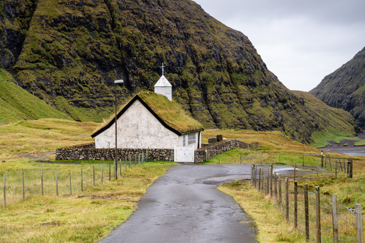 View of the historic Saksun church with a traditional grass roof surrounded by stone walls and lush green hills in the Faroe Islands.