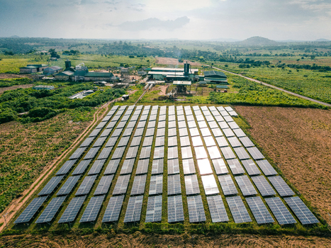 Aerial view of a solar farm with rows of photovoltaic panels industrial buildings in the background and surrounding rural landscape in Ogun State Nigeria.