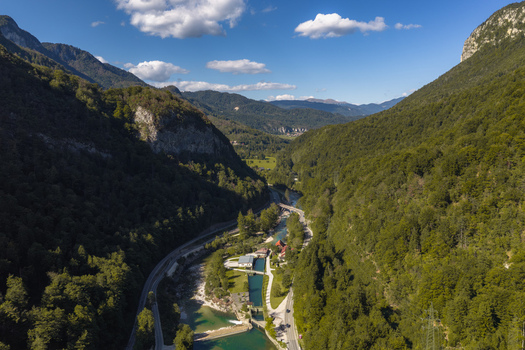 Aerial view of Sava Bohinjka river flowing through a lush green valley surrounded by steep forested mountains under a clear blue sky in Radovljica Slovenia.