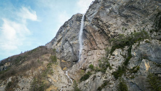 Aerial view of a powerful waterfall cascading down a rugged rock face framed by wispy clouds against a pale blue sky Sallanches Auvergne Rhône Alpes France. Aerial view of a powerful waterfall cascading down a rugged rock face framed by wispy clouds against a pale blue sky Sallanches Auvergne Rhône Alpes France.