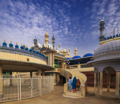 View of the ornate architecture and blue domes of Jamia Masjid Bhong Sharif with people walking up the stairs under a cloudy blue sky in Bhong Punjab Pakistan.