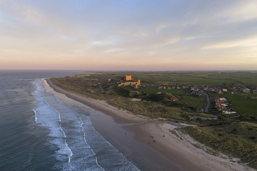 Aerial view of Bamburgh Castle overlooking the sandy beach and North Sea waves under a soft sunset sky Bamburgh England United Kingdom. Aerial view of Bamburgh Castle overlooking the sandy beach and North Sea waves under a soft sunset sky Bamburgh England United Kingdom.