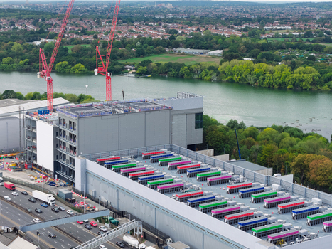 Aerial view of a large data center construction site featuring red cranes and rows of colorful rooftop cooling units situated next to a river and a busy highway in the United Kingdom.