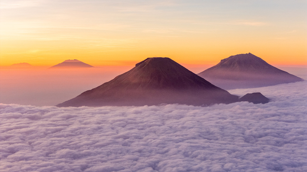 Aerial view of Mount Bismo and surrounding volcanic peaks rising above a thick sea of clouds during a vibrant golden hour sunrise in Kecamatan Kejajar Jawa Tengah Indonesia.