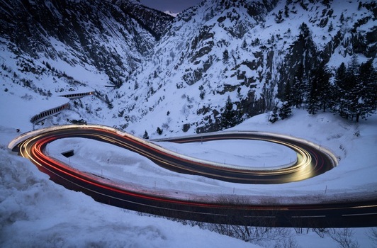 View of light trails on the winding Sankt Gotthard Pass road surrounded by snow covered mountains at dusk in Airolo Ticino Switzerland.