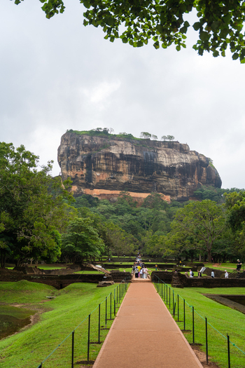 View of Sigiriya Rock Fortress with tourists walking along a central path through lush green gardens under an overcast sky Sigiriya Central Province Sri Lanka. View of Sigiriya Rock Fortress with tourists walking along a central path through lush green gardens under an overcast sky Sigiriya Central Province Sri Lanka.