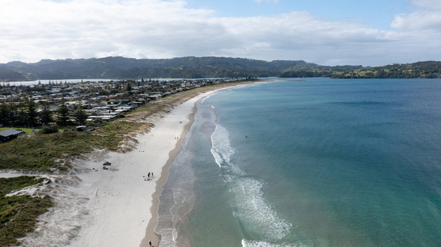Aerial view of Omaha Beach with white sand turquoise water coastal residential area and rolling hills in the background Omaha Auckland New Zealand.