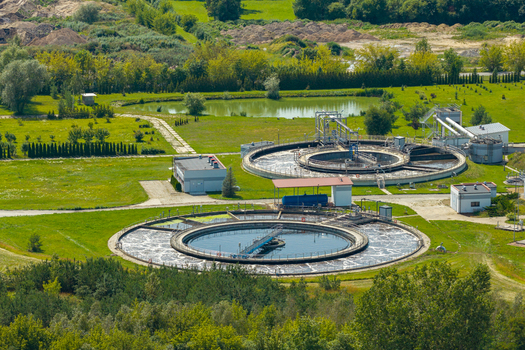 Aerial view of a wastewater treatment plant with circular sedimentation tanks surrounded by green fields and trees in Poland.