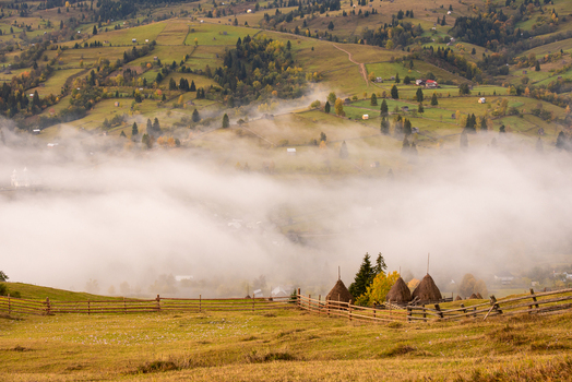 View of traditional haystacks in a misty rolling rural landscape with wooden fences and autumn foliage in Moldovita Suceava County Romania. View of traditional haystacks in a misty rolling rural landscape with wooden fences and autumn foliage in Moldovita Suceava County Romania.