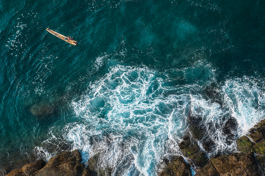 Aerial view of a small wooden boat floating on turquoise sea water near rocky cliffs with crashing waves Kovalam Kerala India. Aerial view of a small wooden boat floating on turquoise sea water near rocky cliffs with crashing waves Kovalam Kerala India.