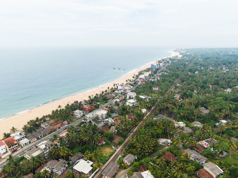 Aerial view of the sandy coastline and turquoise ocean with palm trees and houses along the road and railway track in Dodanduwa Southern Province Sri Lanka. Aerial view of the sandy coastline and turquoise ocean with palm trees and houses along the road and railway track in Dodanduwa Southern Province Sri Lanka.