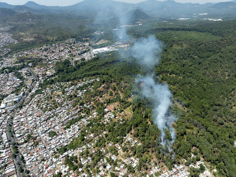 Aerial view of a wildfire burning in the forest near the urban outskirts with thick smoke rising above the trees and residential buildings in Uruapan Michoacan Mexico.