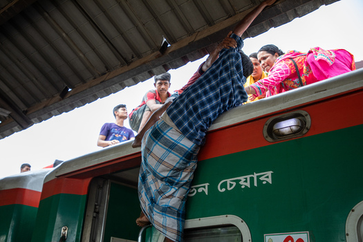 Dhaka Bangladesh 14 June 2018 View of people climbing onto the roof of a train at Airport Railway Station with a man in a lungi hanging from the side of a green carriage. Dhaka Bangladesh 14 June 2018 View of people climbing onto the roof of a train at Airport Railway Station with a man in a lungi hanging from the side of a green carriage.