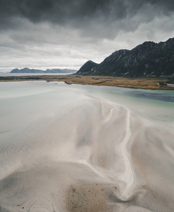 Aerial view of the sandy tidal flats and turquoise waters along Midnattsolveien with rugged mountains under an overcast sky in Laukvik Nordland Norway. Aerial view of the sandy tidal flats and turquoise waters along Midnattsolveien with rugged mountains under an overcast sky in Laukvik Nordland Norway.
