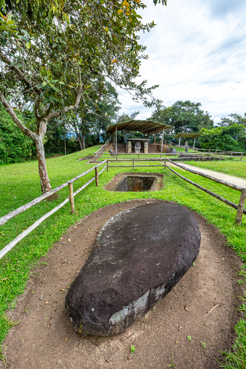 View of the ancient stone monolith at San Agustin Archaeological Park surrounded by lush green grass trees and wooden fencing in San Agustin Huila Colombia.