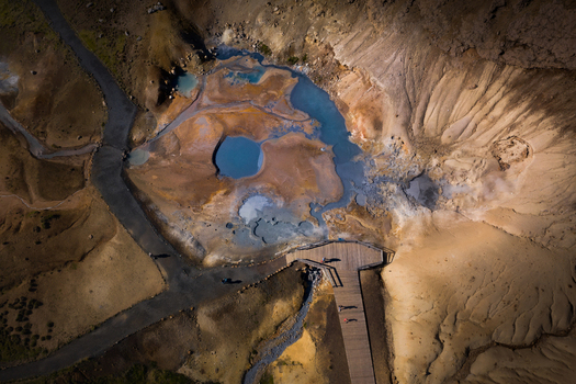 Aerial view of geothermal pools with blue water and steaming vents surrounded by mineral rich soil and a wooden boardwalk in Iceland.