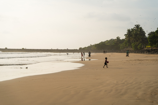 View of people walking along the sandy shoreline and a child playing with a ball near the historic fortifications and palm trees in Galle Southern Province Sri Lanka.